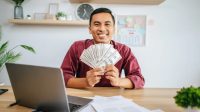 seorang pegawai lelaki memegang uang dengan laptop dan meja di depannya